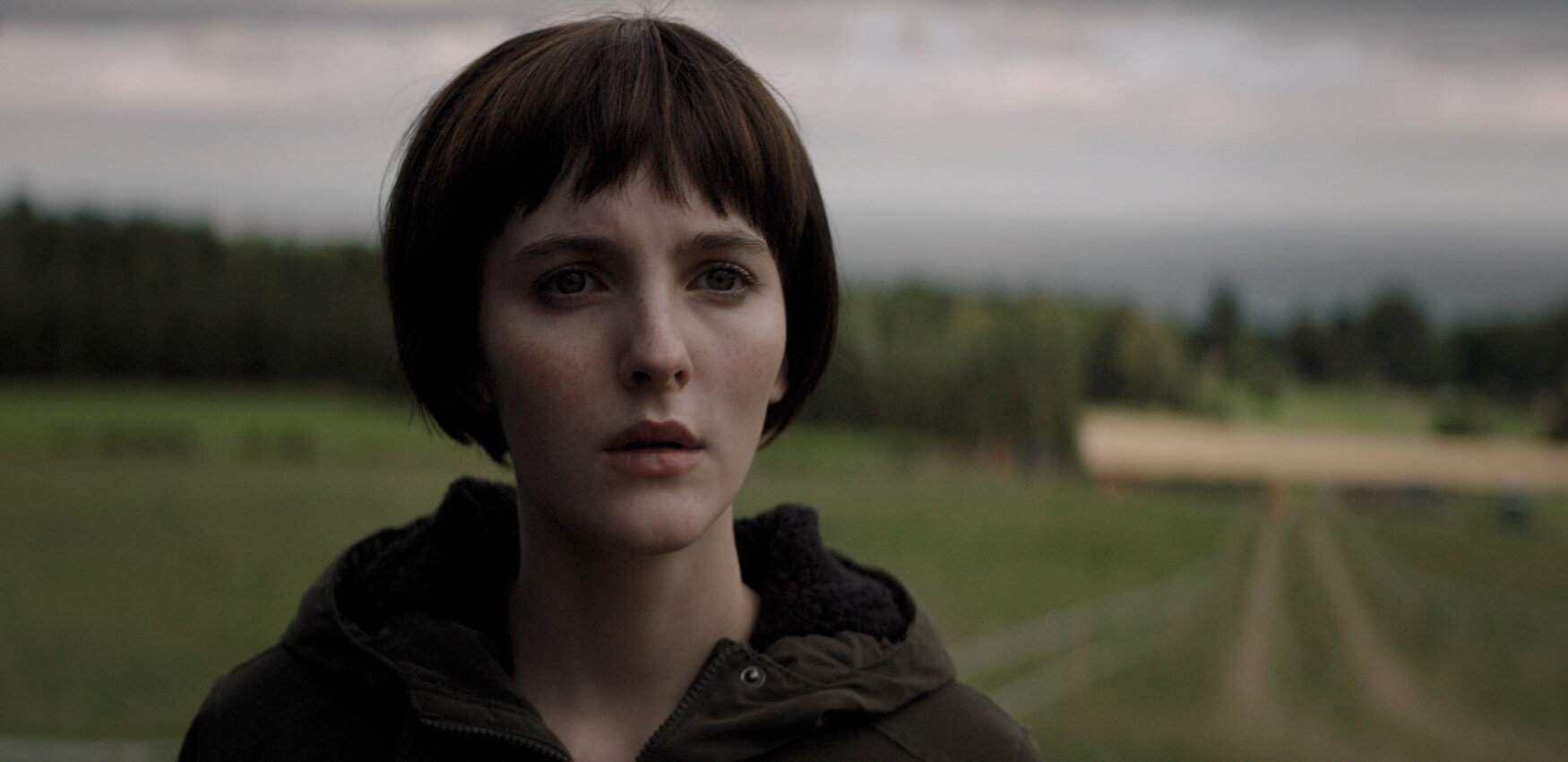 a pale white woman with a black bob stands in a field. a scene from the film ROSE PLAYS JULIE. hear head is close up and in focus, she looks out confused, as the green field and overcast sky are blurred in the background.