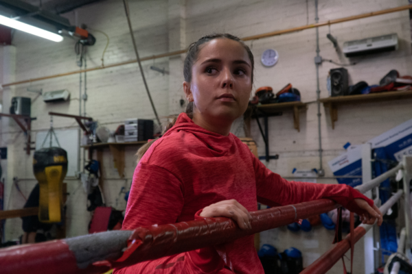 a girl looks out wistfully from a. boxing ring. she wears a pink sports top and leans over red rope.