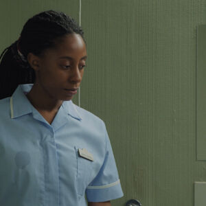 A nurse stands in a bathroom of a nursing home. the film still frames from her waist up, we see her looking down with an engaged and caring expression