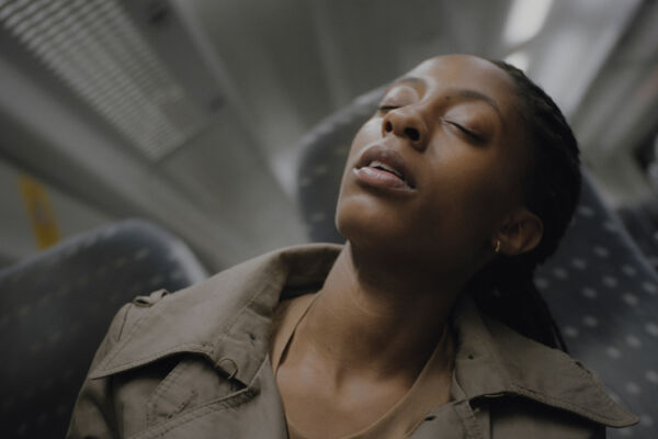 film still. close up of a woman sat on a train, shot at an angle she seems to be falling off. her chair. to sleep.