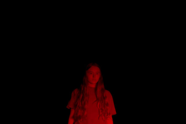 a girl stands in the middle of a black night scene. she is illuminated in red, looking down intently.