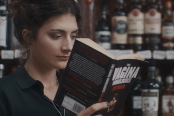 a woman reads a copy of 'the vagina monologues'. she reads intently. despite her surroundings, the alcohol aisle in an off licence