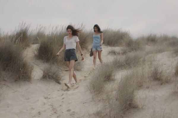 two girls walk across sand dunes. it is a grey day and the wind blows their hair at the same angle as the bushes of dry shrubs.