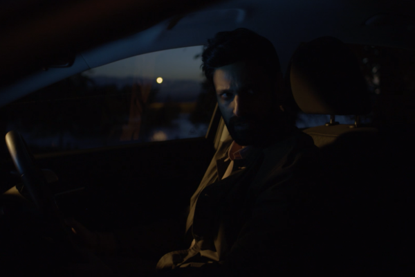 a dark image, of a man siting in the drivers seat of the car. the camera is placed in passenger side, he is leaning towards.