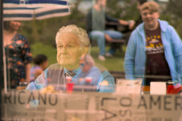 an old man looking out a large window from a cafe table. a young boys face reflected in the window - they smile at eachother