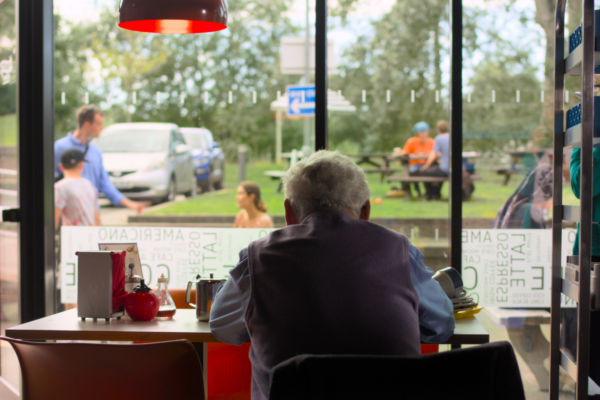 wide shot of the back of an old man sat at a table at a service station - looking out from inside at people arguing in the outdoor space.
