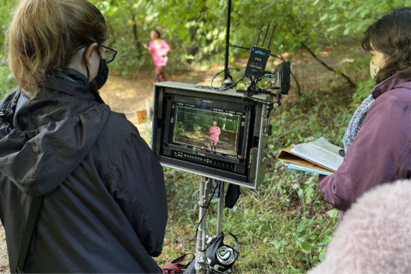 A person stands looking at a monitor on a film set. They are under an umbrella, stood in a waterproof in the woods. The image is green and luscious as they are in the woods. on the monitor, and visible in the background of the image another person is running in pink.