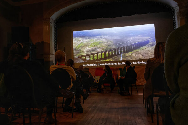 a large cinema screen looms above an audiences. the screen shows an aerial shot of a green field, with an aqueduct going through