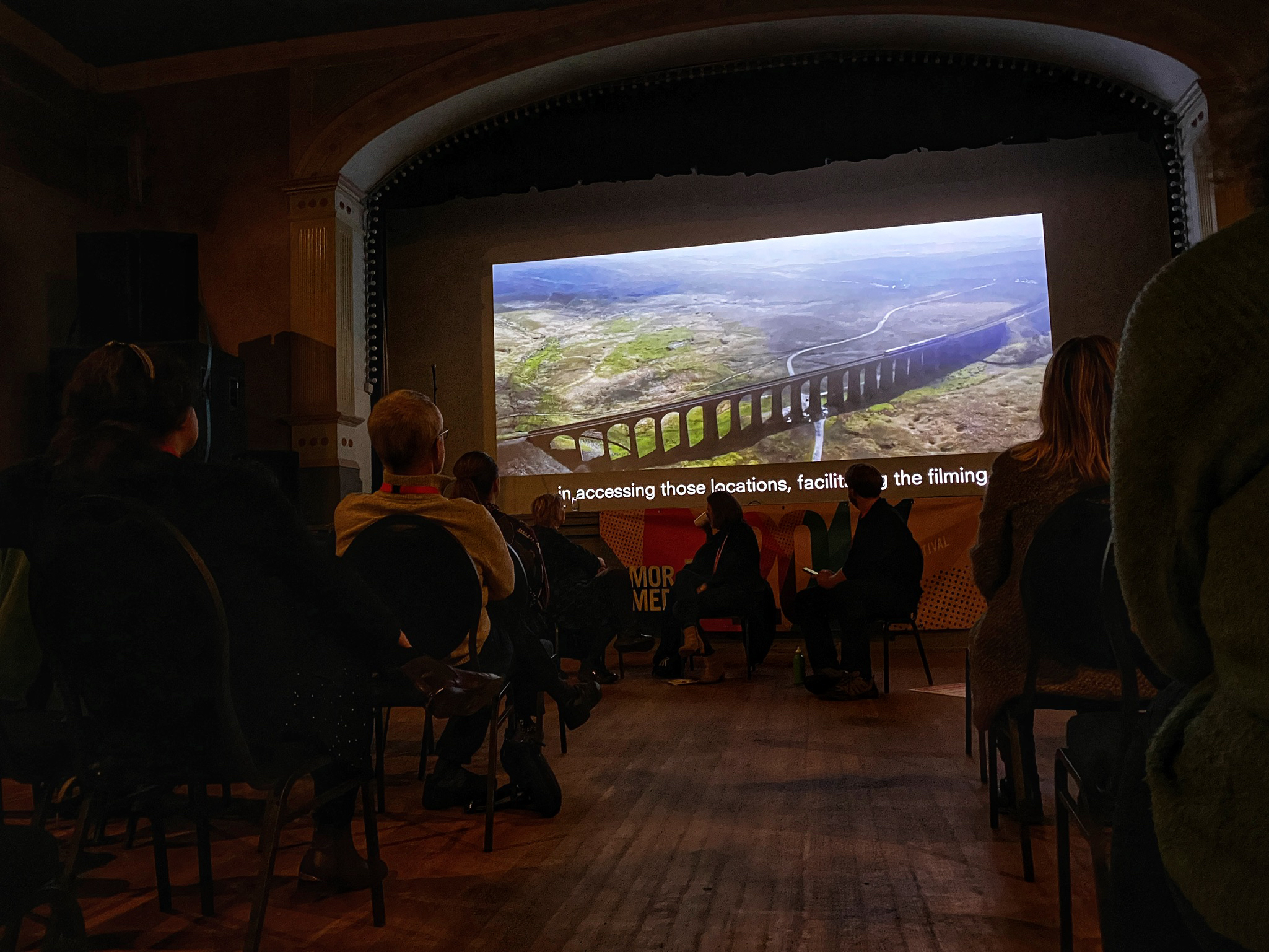 a large cinema screen looms above an audiences. the screen shows an aerial shot of a green field, with an aqueduct going through
