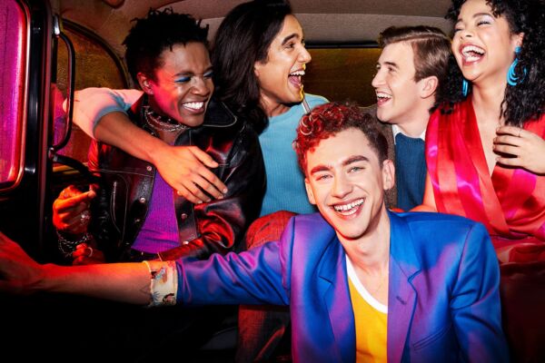A press photo from Channel 4 TV Drama series 'IT'S A SIN'. A group of people are sat in the back of a taxi laughing and smiling, sat on top of each other. They are dressed colourfully, and a pink light illuminates them.
