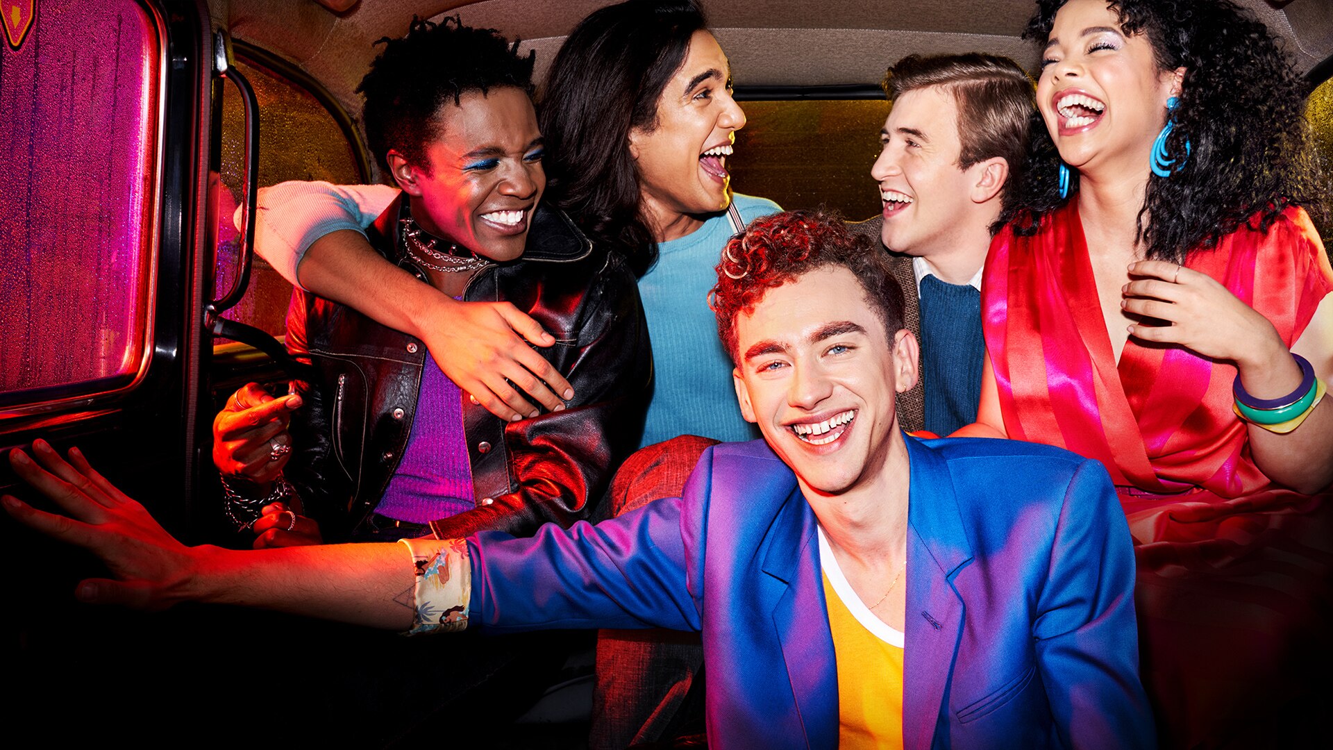 A press photo from Channel 4 TV Drama series 'IT'S A SIN'. A group of people are sat in the back of a taxi laughing and smiling, sat on top of each other. They are dressed colourfully, and a pink light illuminates them.