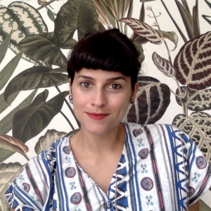 headshot of Nadia Attia. She is a woman with a fringe of short dark hair. she wears red lipstick and a patterned top. she sits infront of a background of a leafy print