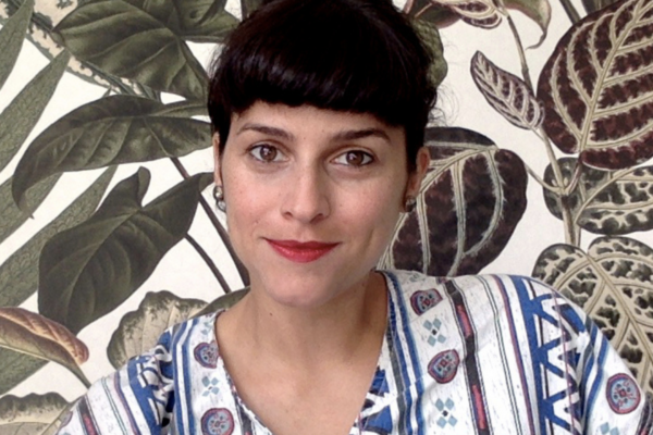 headshot of Nadia Attia. She is a woman with a fringe of short dark hair. she wears red lipstick and a patterned top. she sits infront of a background of a leafy print