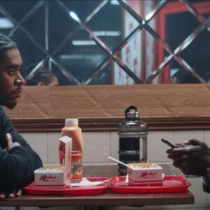 no more wings film still. two men sit opposite each other in a chicken shop. food sits between them. one on the right stares down at his phone, the other sits across, looking frustrated with his companion.