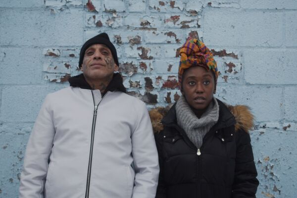two people stand together infront of a distressed blue brick wall. on the left, a man with face tattoos wears a white jacket. a woman stands close to his right, wearing a fluffy black coat and Ghanian head wrap. they look exasperated