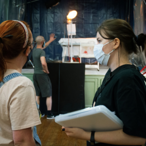 Two women stand on set, looking over a clipboard and a monitor. They are on a filmmaking team, and stand in the centre of the image, talking to each other and framed from the waist up. A person in the back stands and moves a light.