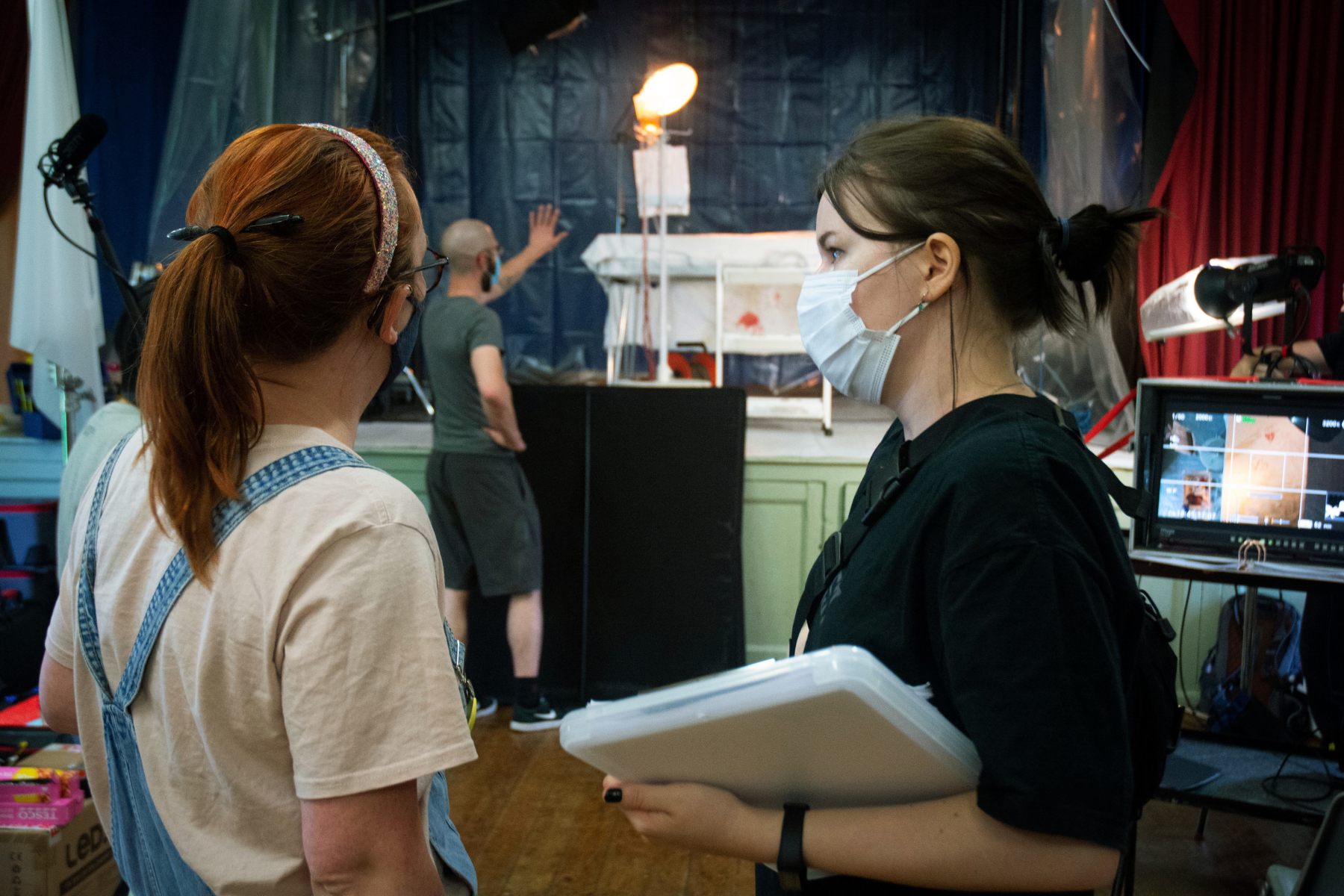 Two women stand on set, looking over a clipboard and a monitor. They are on a filmmaking team, and stand in the centre of the image, talking to each other and framed from the waist up. A person in the back stands and moves a light.