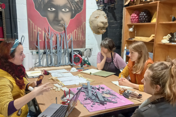 Costume design meeting, four people sit around a table cluttered with animation tools, scraps, sculptures, and figurines