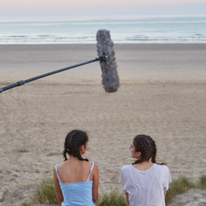 two girls sit on a beach at sunrise, and a boom mic hovers over their heads and the horizon