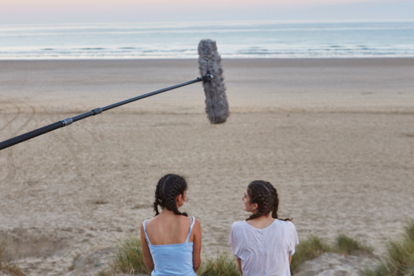 two girls sit on a beach at sunrise, and a boom mic hovers over their heads and the horizon