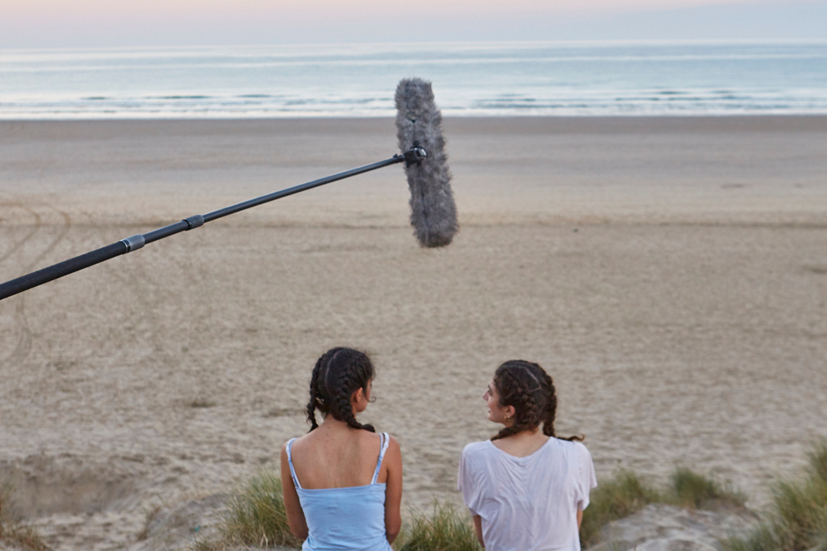 two girls sit on a beach at sunrise, and a boom mic hovers over their heads and the horizon