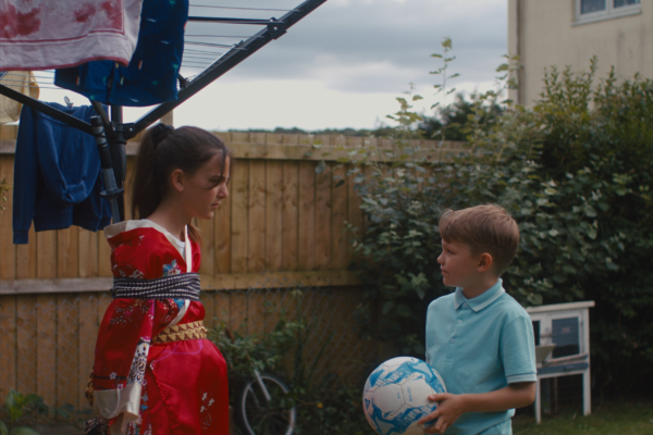 two children stand in their garden. one is tied to a washing line pole, and the other looks at them while holding a football. it is a nice moment on an overcast day