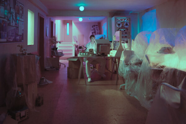 Wide image of a the main character sat at a desk in an 80s style lab, with blue and pink lighting