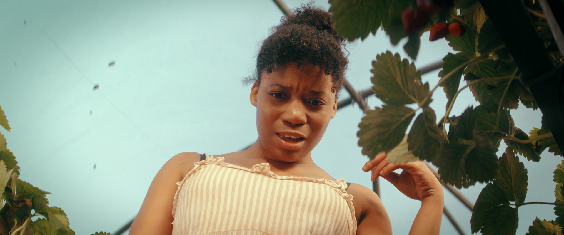 A black woman looks down at the camera, in a yellow vest, with foliage behind her