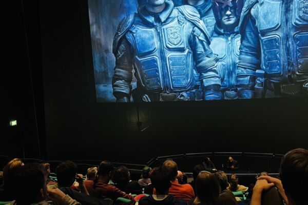 A talk in the former Bristol IMAX with two speakers at a table in front of the screen which is displaying an image from the film 'Dredd'. The audience are in the foreground listening to the speakers.
