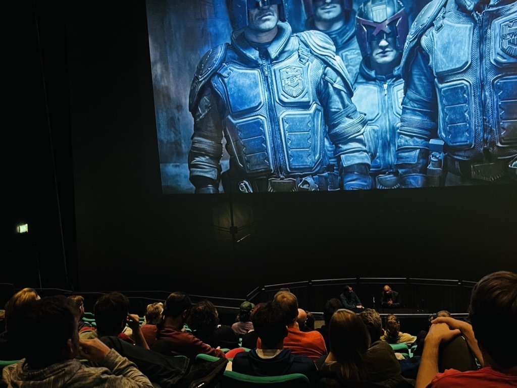 A talk in the former Bristol IMAX with two speakers at a table in front of the screen which is displaying an image from the film 'Dredd'. The audience are in the foreground listening to the speakers.