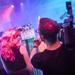 A pink, blue and purple-lit room with a camera man, clapperboard and a female actor in a long pink wig.