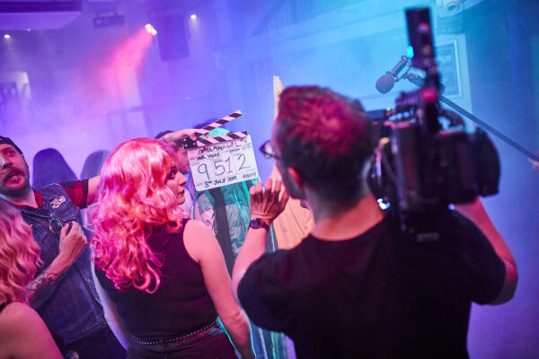 A pink, blue and purple-lit room with a camera man, clapperboard and a female actor in a long pink wig.