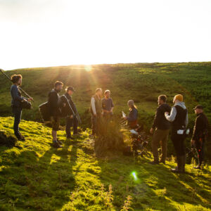 Behind the scenes of a film shoot in a green landscape with a film crew and a sunset behind them