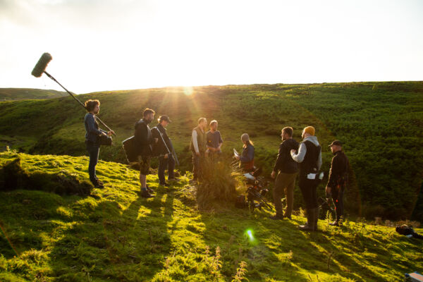 Behind the scenes of a film shoot in a green landscape with a film crew and a sunset behind them