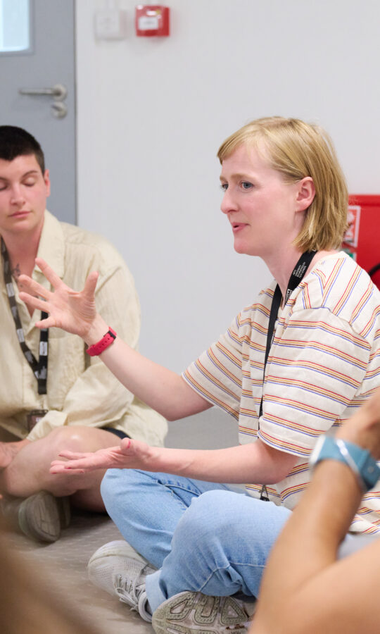 Em Smith, a blonde woman with a short bob, wearing a pale yellow and white stripy t-shirt, gestures expressively while others listen attentively.