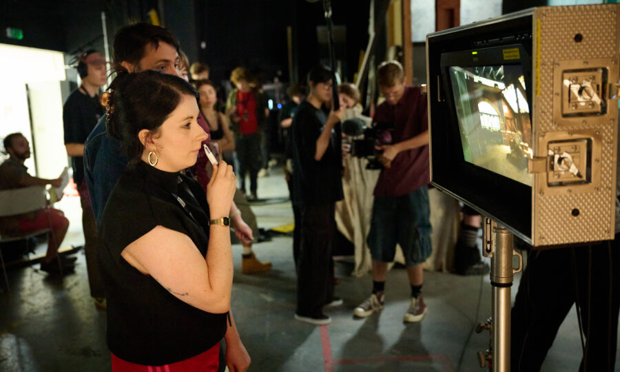 A woman attentively observes a monitor on a busy film set, surrounded by crew members and equipment. The scene conveys focus and collaboration.