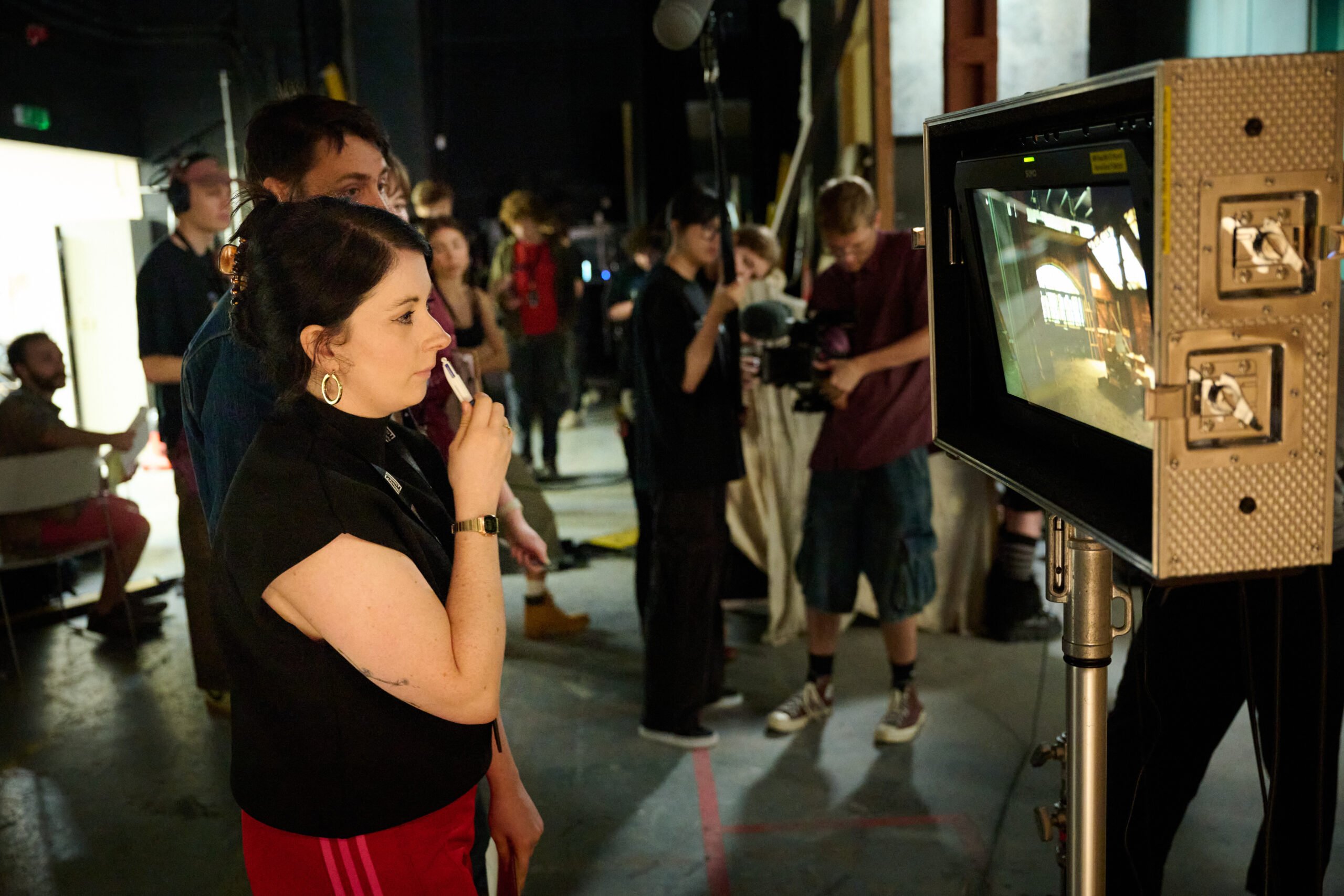 A woman attentively observes a monitor on a busy film set, surrounded by crew members and equipment. The scene conveys focus and collaboration.