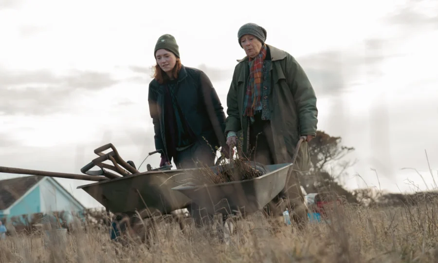 Two women in warm clothing push a wheelbarrow filled with gardening tools across a field. The sky is overcast, creating a calm, focused atmosphere.