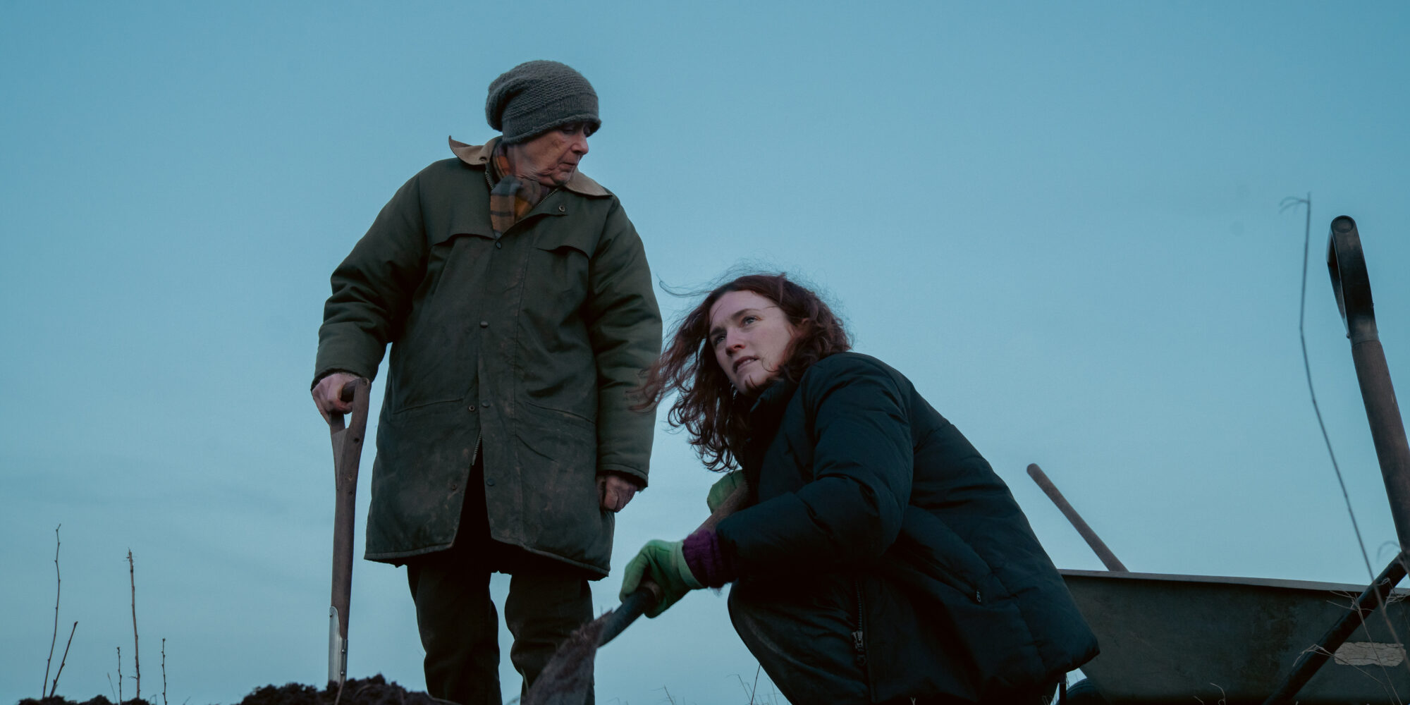 Two women in winter clothes work outdoors with shovels; one stands and the other kneels by a wheelbarrow against a dusk sky. The mood is somber.