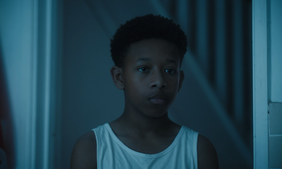 Young boy in a white tank top stands in a dimly lit room, casting a thoughtful gaze. Soft blue lighting and shadows create a somber, contemplative mood.