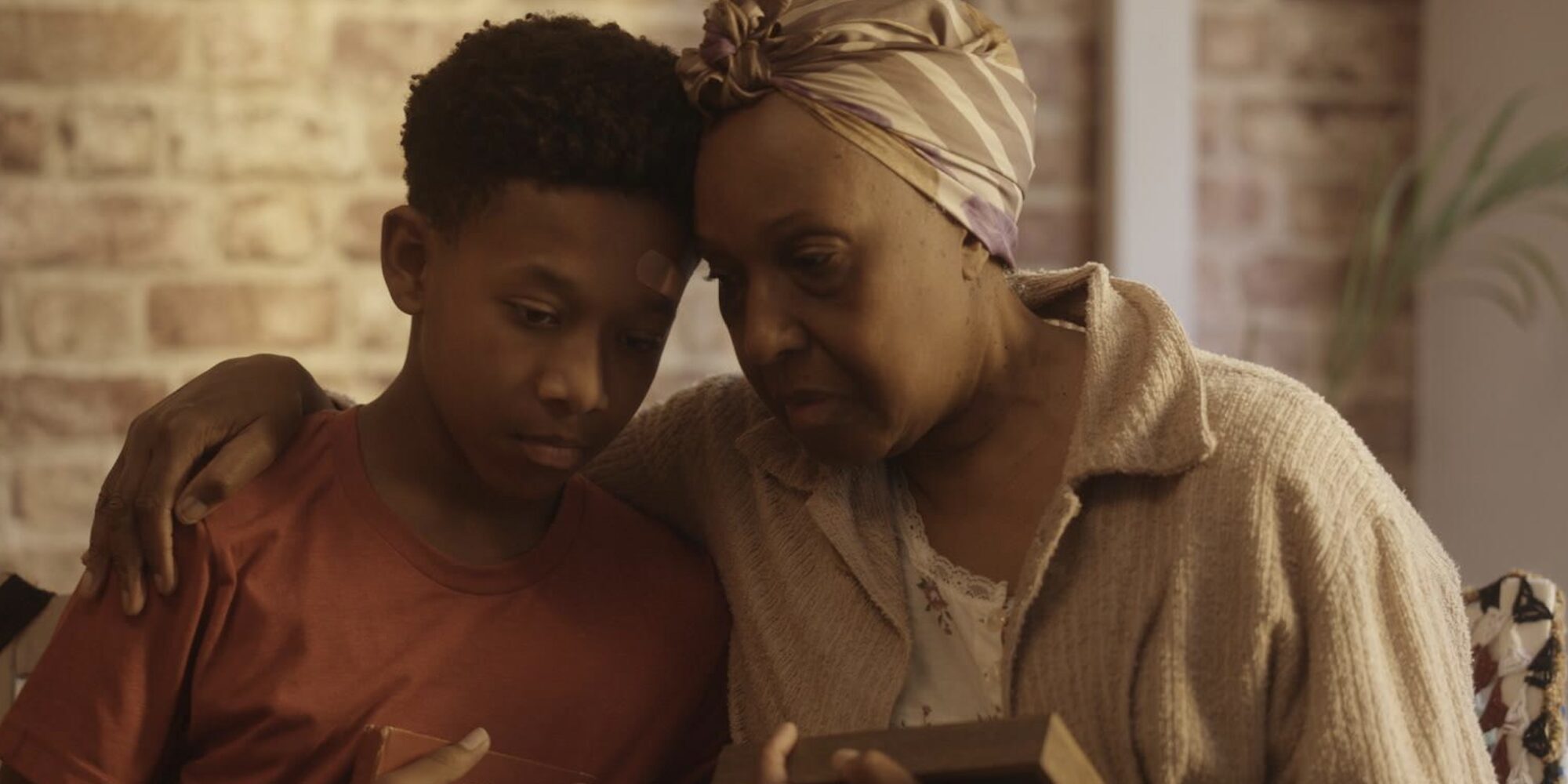 An elderly woman and a young boy share an emotional moment, sitting closely with arms around each other. They look somber, holding a framed photo.