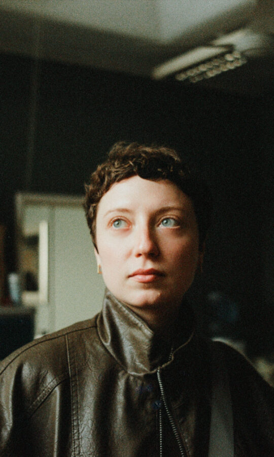 Thea Gajic headshot. A woman with short hair looks thoughtfully to the side in a dimly lit room. They're wearing a leather jacket, surrounded by shelves and boxes.
