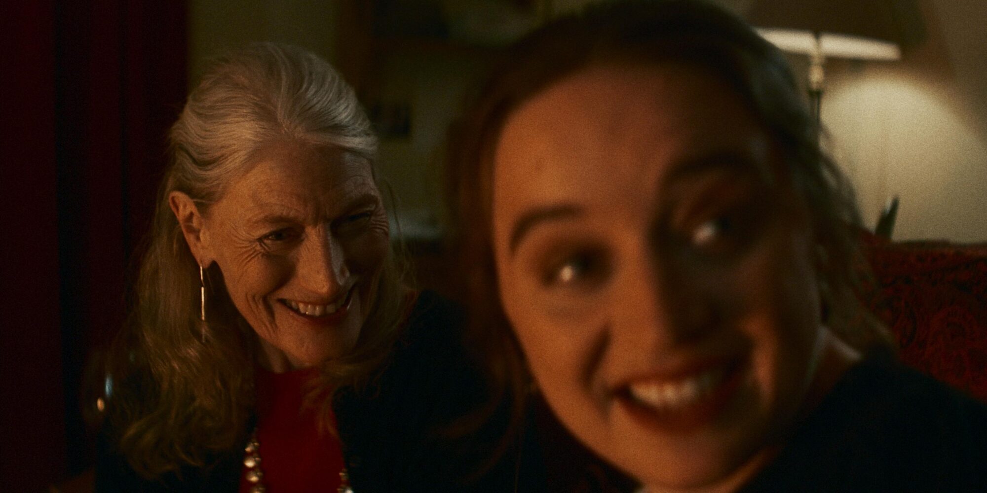 Two women in a warmly lit room. The older woman with grey hair and earrings smiles warmly, while the younger woman laughs exuberantly.