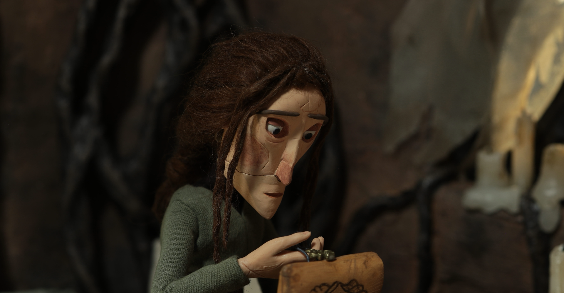 A claymation character with long hair, wearing a green sweater, opens a small engraved wooden chest.