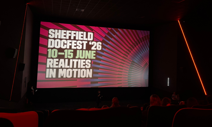 A large cinema screen illuminates a dark cinema featuring a presentation slide reading: Sheffield DocFest '26, 10 - 15 June, Realities In Motion