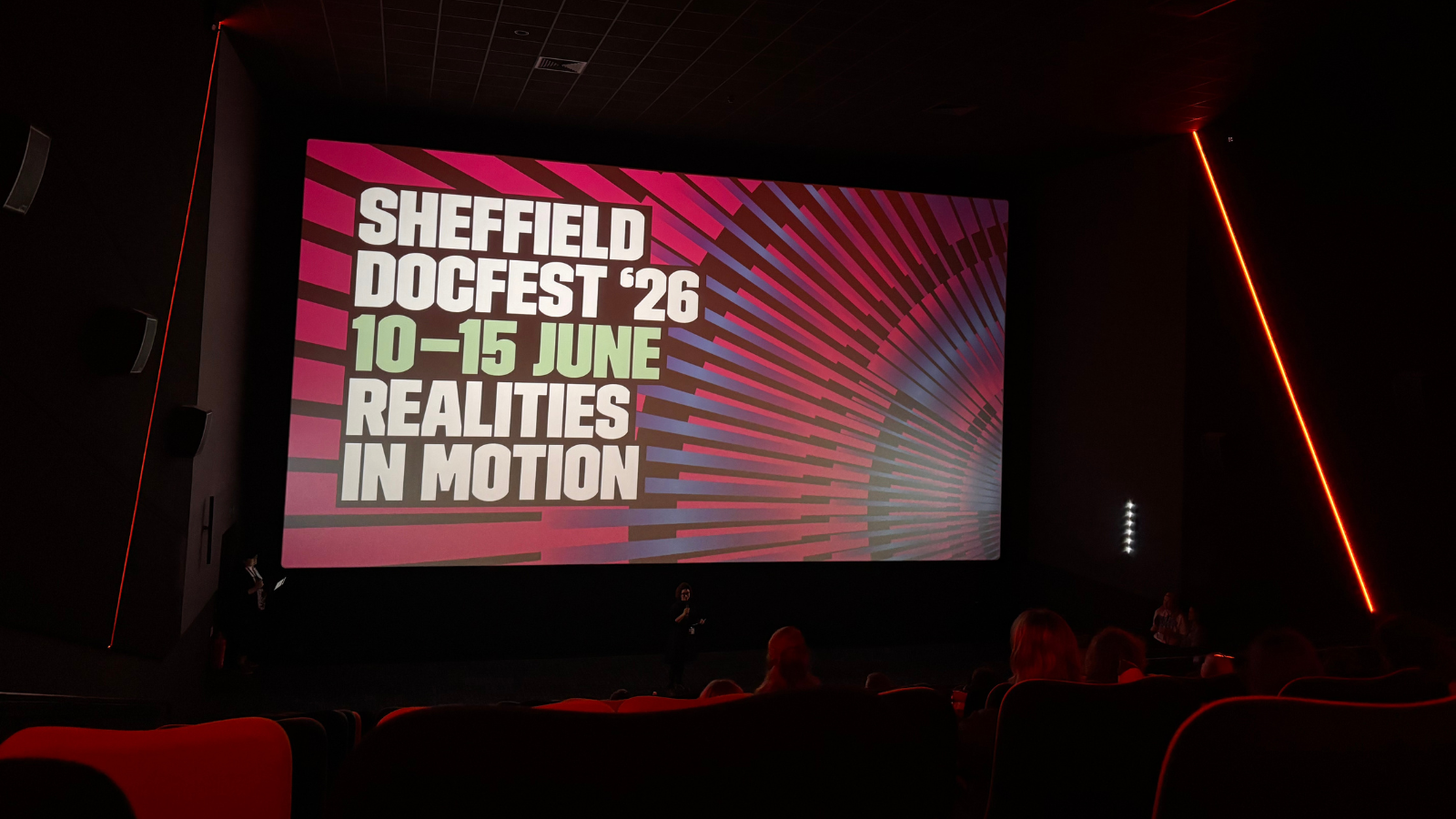 A large cinema screen illuminates a dark cinema featuring a presentation slide reading: Sheffield DocFest '26, 10 - 15 June, Realities In Motion
