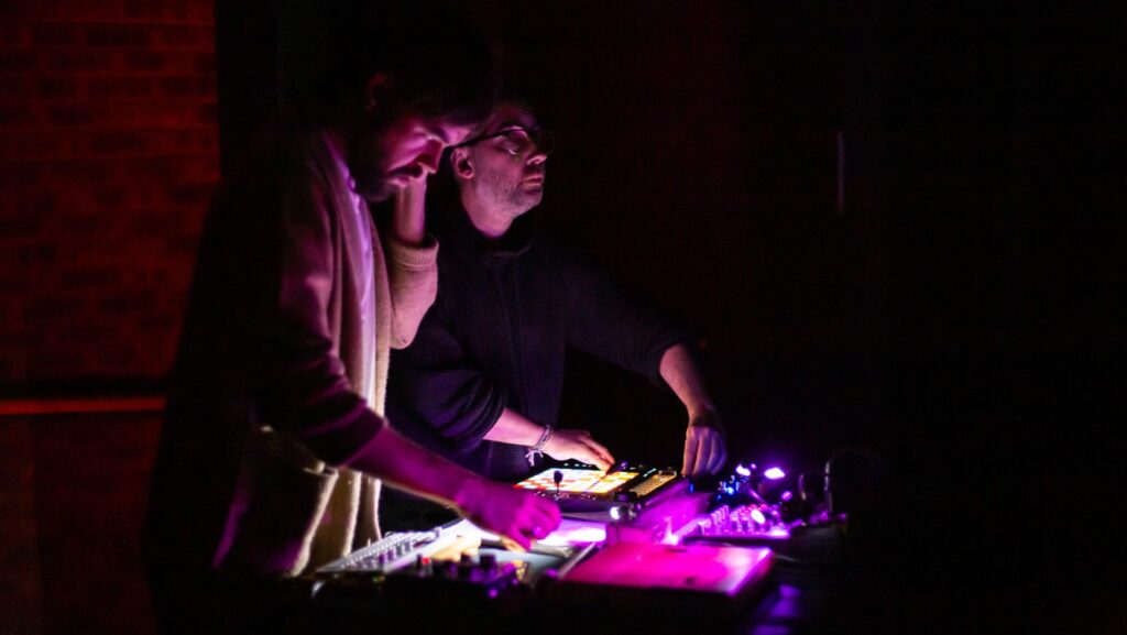 Two people stand in a dark control room, illuminated by a glowing purple lighting console in front of them.