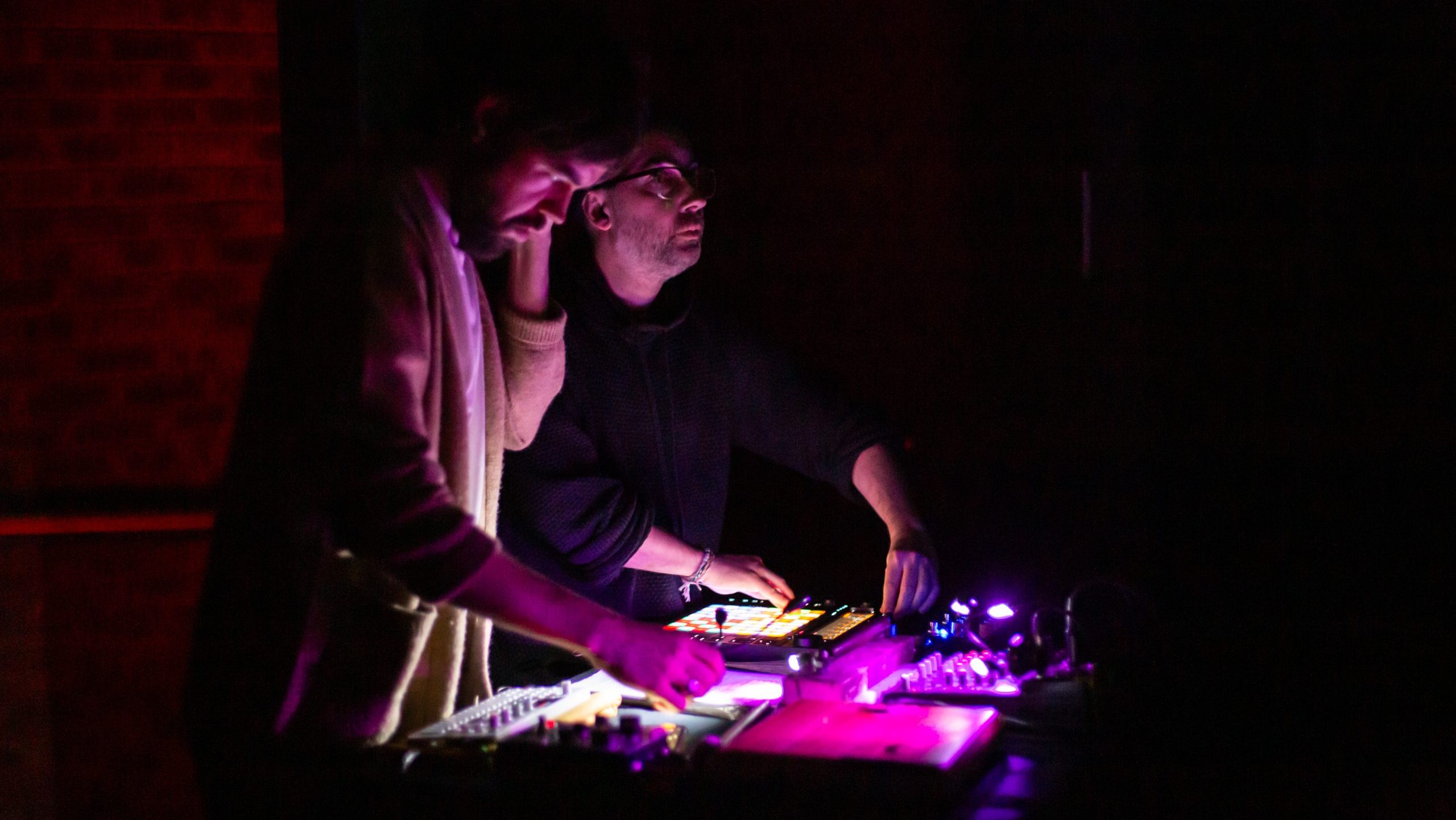 Two people stand in a dark control room, illuminated by a glowing purple lighting console in front of them.
