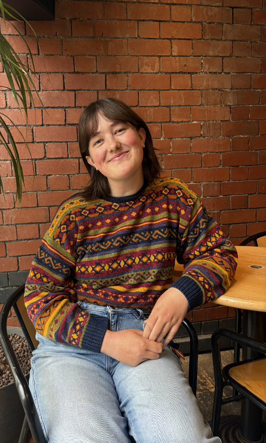 A woman in a colourful patterned jumper sits at a wooden table in front of a red brick wall.