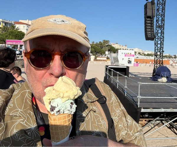 A man wearing sunglasses and a cap holds an ice cream cone at an outdoor event near a stage.